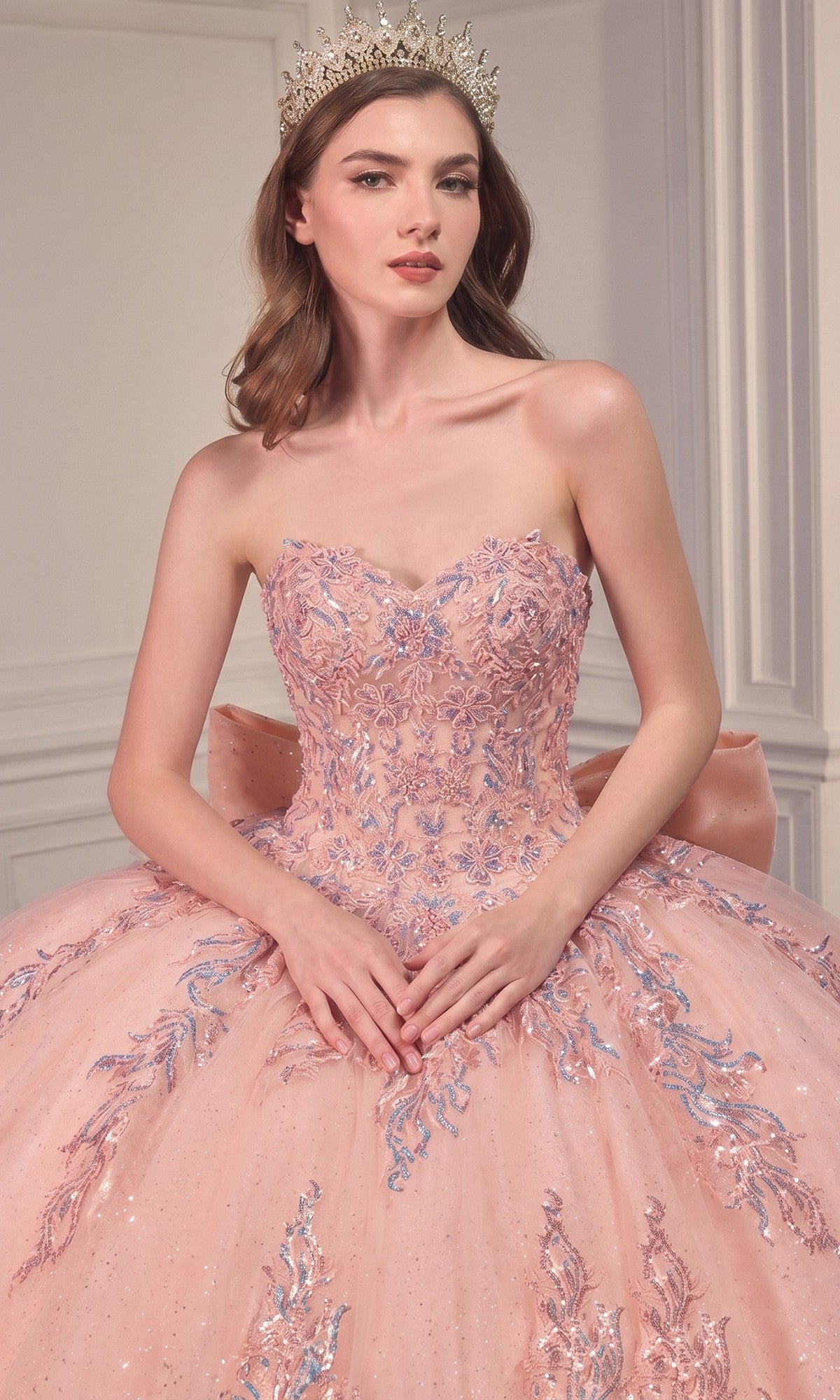 A woman poses indoors against a white paneled wall wearing the Ladivine Strapless Quinceanera Dress 15715, a pink gown featuring an embellished ball gown skirt and paired with a jeweled crown.